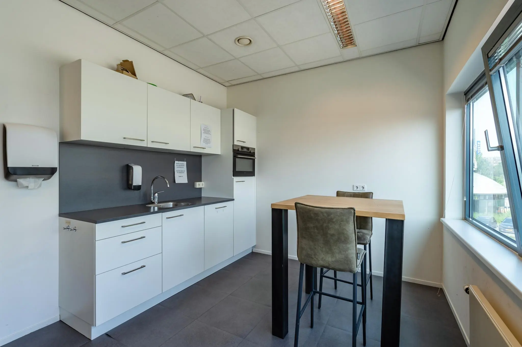 Moderne kantoorkeuken met witte kasten, een hoge eettafel en zicht op Rivium Boulevard.